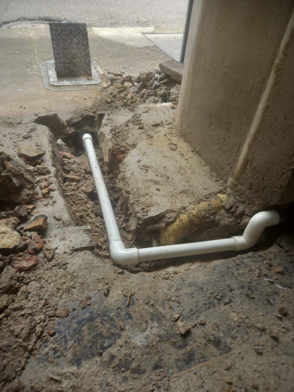 Reparación de tubería de PVC blanca en una zanja de tierra junto a un muro y un pilar de hormigón.