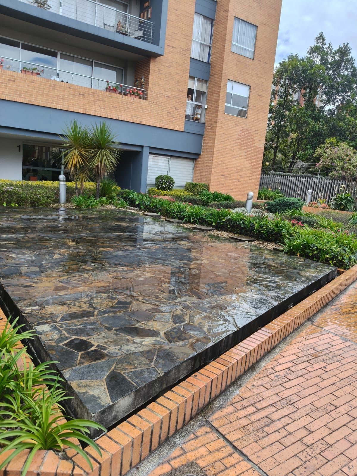 Patio con terraza de piedra húmeda, bordes de ladrillo, zonas verdes ajardinadas y un moderno edificio de apartamentos.