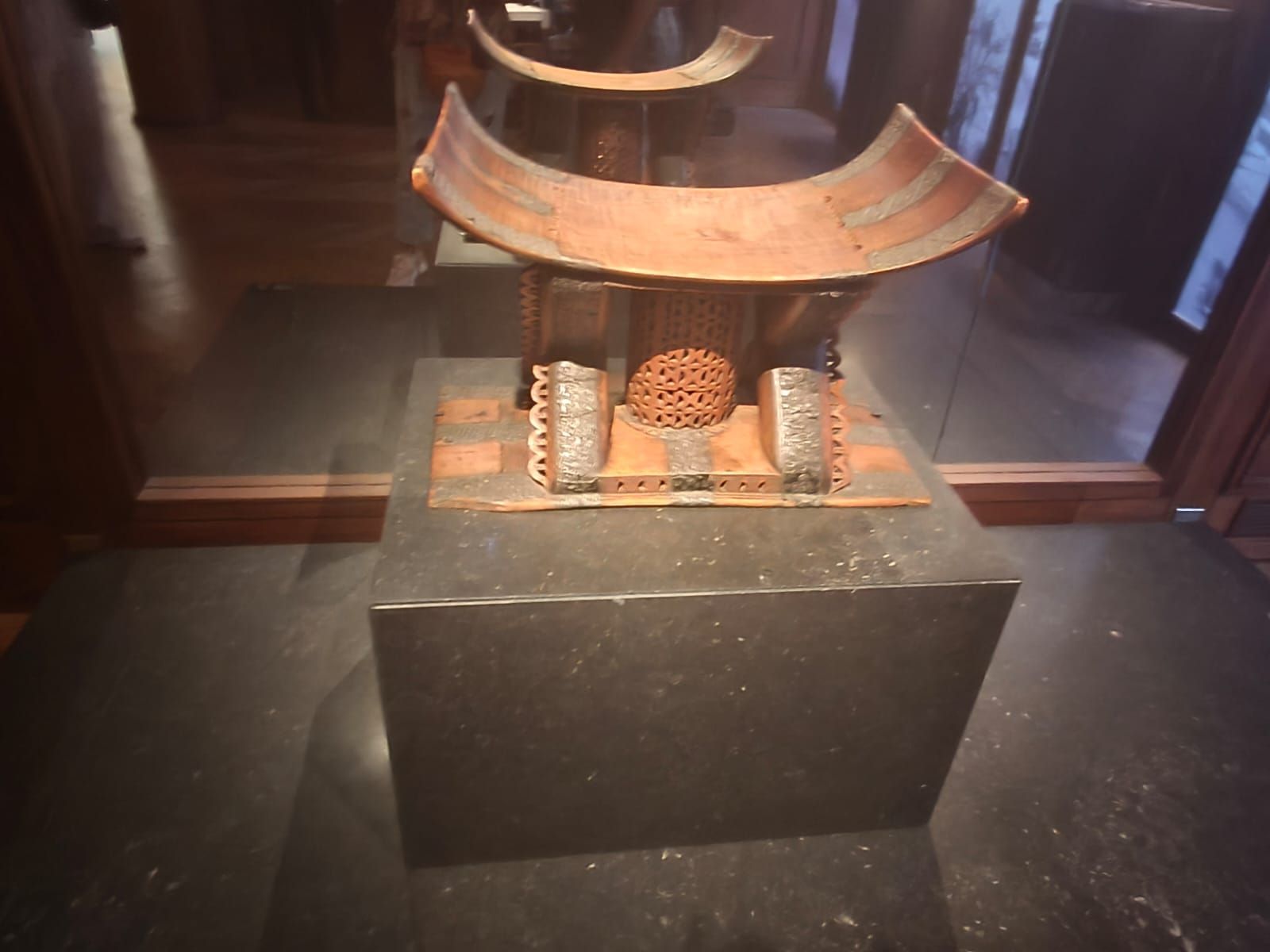 Pequeña escultura de bronce con forma de templo y techo curvo sobre un pedestal negro, expuesta en una vitrina de museo.