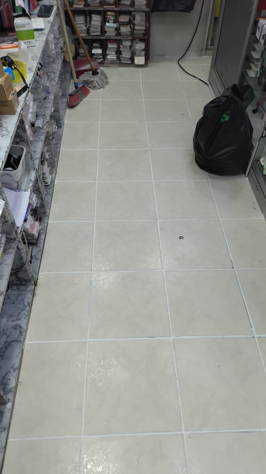 Pasillo de tienda con suelo de baldosas beige, estanterías a ambos lados y una bolsa negra cerca de la pared derecha.