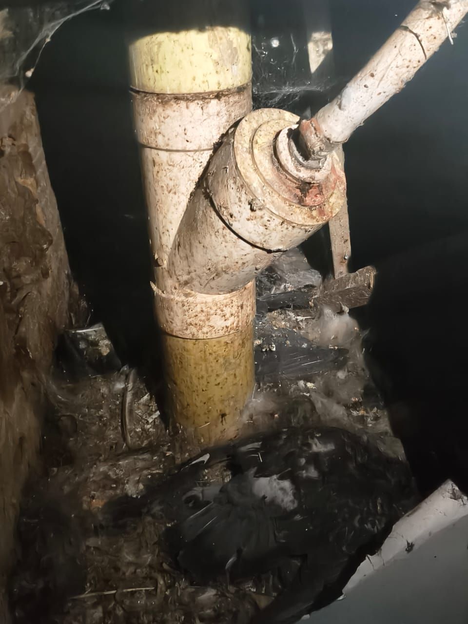 Tuberías industriales oxidadas y una válvula en una zona de servicios oscura y sucia, con agua o lodo debajo.