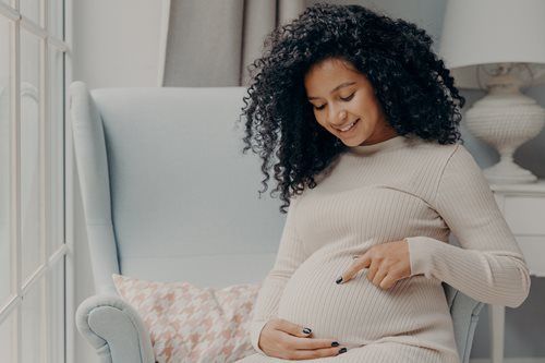 A pregnant person sits in an armchair, smiling down at their stomach and touching it with their finger.