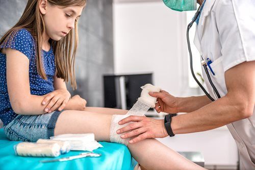 A medical professional wearing a mask and white coat wraps a bandage around a patient’s leg in a clinical setting.