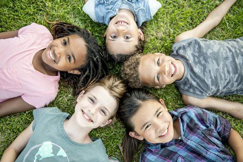 Five smiling friends lying in a circle on grass, looking up at the camera.