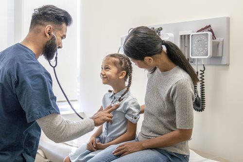 A healthcare worker uses a stethoscope to examine a child sitting on an exam table while an adult supports them.