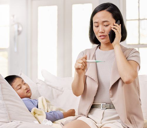 A person in a neutral-toned outfit sits on a couch, holding a thermometer while talking on a phone, with a child nearby.