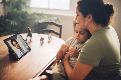 Woman and child sitting at a table, watching a tablet with a video call on screen
