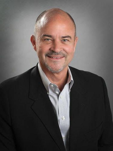 Bald man smiles, wearing a black blazer over a striped shirt. Gray background.