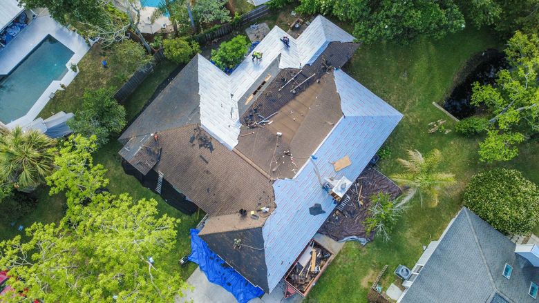 Aerial view of a house with a damaged roof, surrounded by trees and neighboring homes.