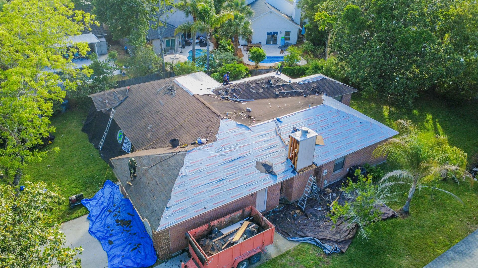 Aerial view of a house roof under construction with workers and a red dumpster in a green suburban yard.