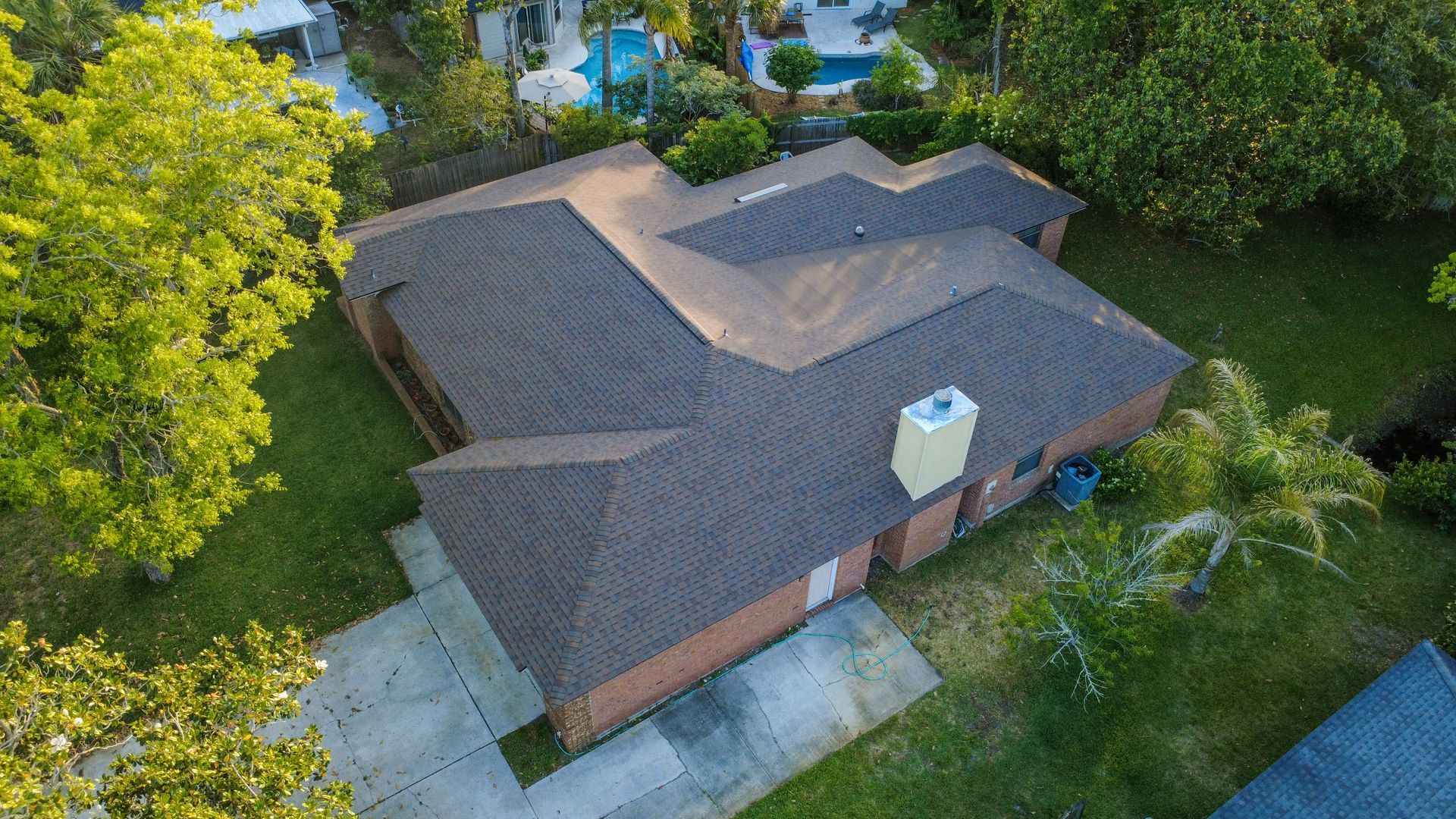 Aerial view of a brown-roofed house with a driveway, surrounded by green trees and lawn.