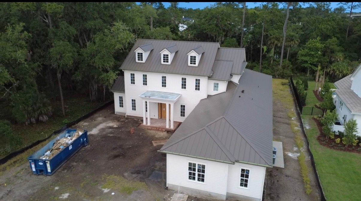 Aerial view of a large white house under construction with a blue dumpster and surrounding trees