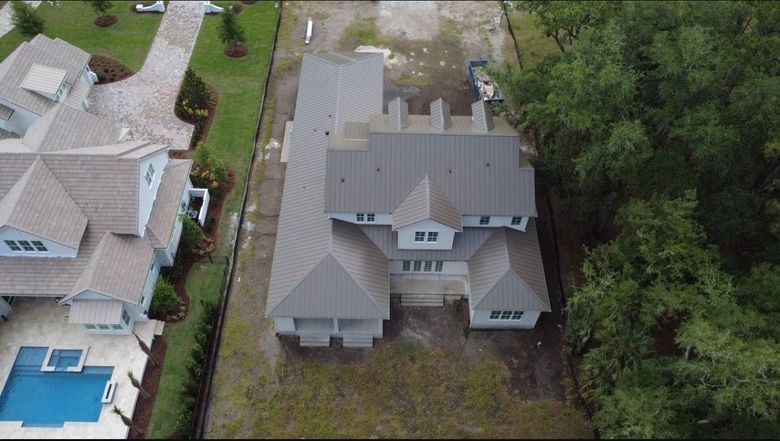 Aerial view of a gray house with a metal roof, driveway, and trees beside neighboring homes.