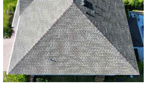 Aerial view of a gray shingled house roof with a steep gable and surrounding trees.