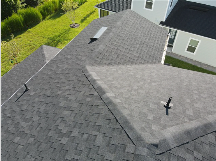 Aerial view of gray shingled house roofs and a dormer beside a green lawn and white siding.