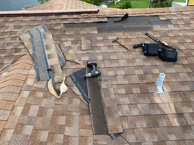 Roof with shingle repair materials and tools laid out on a brown asphalt shingle surface.