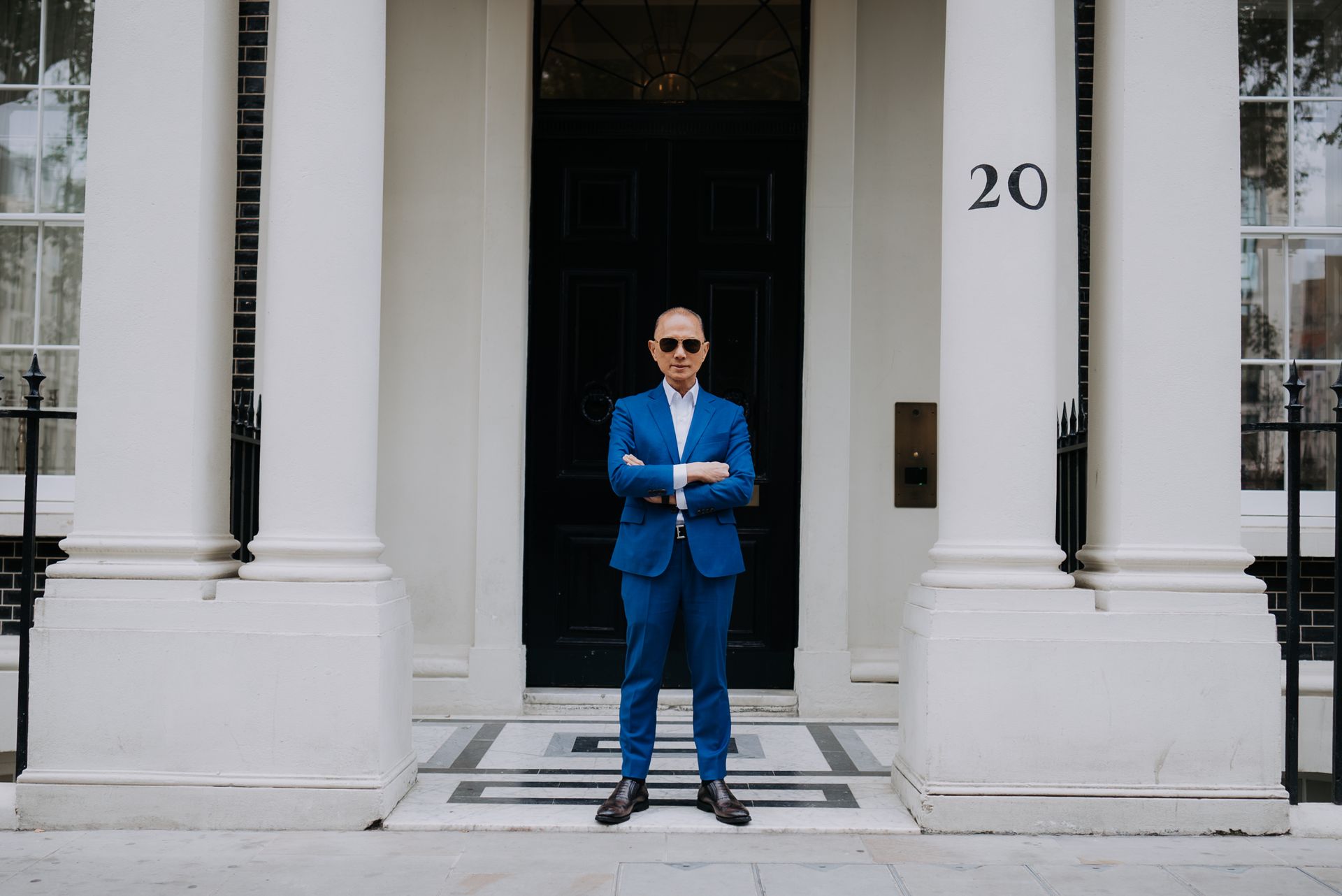 Jimmy Choo in blue suit stands in front of a white building with the number 20 on the door.