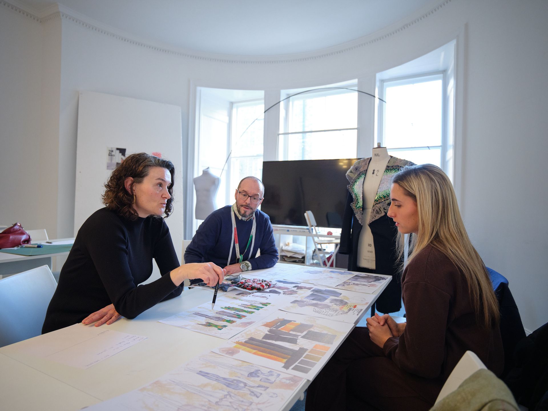 Three people in a bright room reviewing a large blueprint on a table. One person points, others look on.