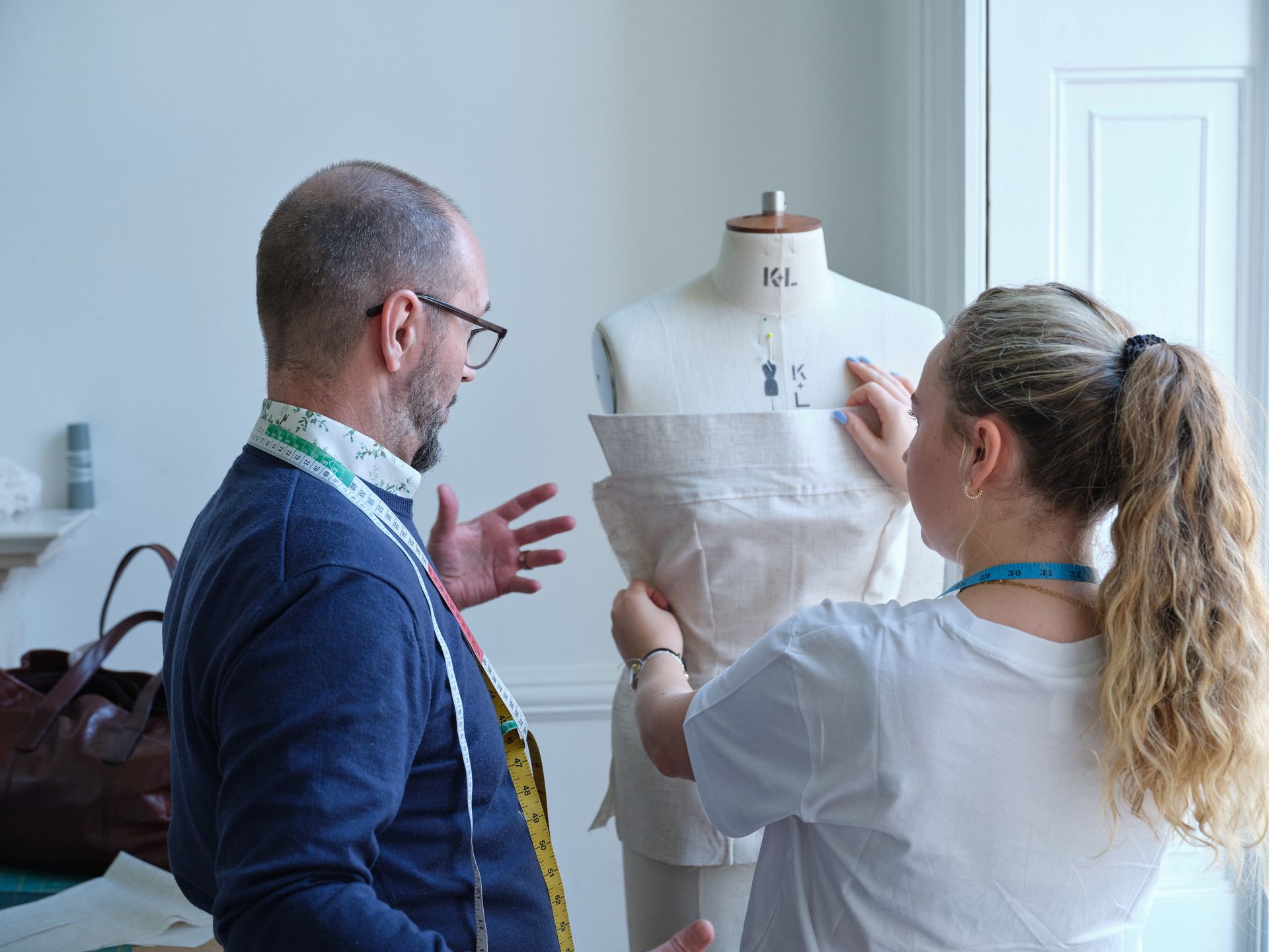 A man and a woman work on garment construction around a dress form, indoors.
