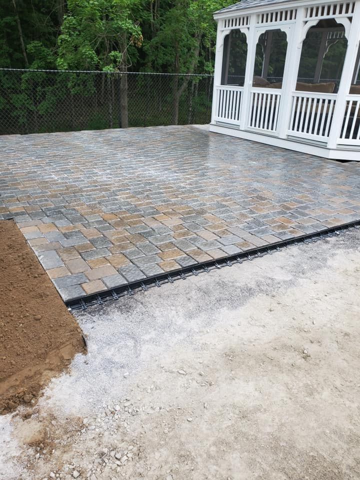 A gray and brown brick patio under construction, bordering a white gazebo and an unpaved, sandy area.