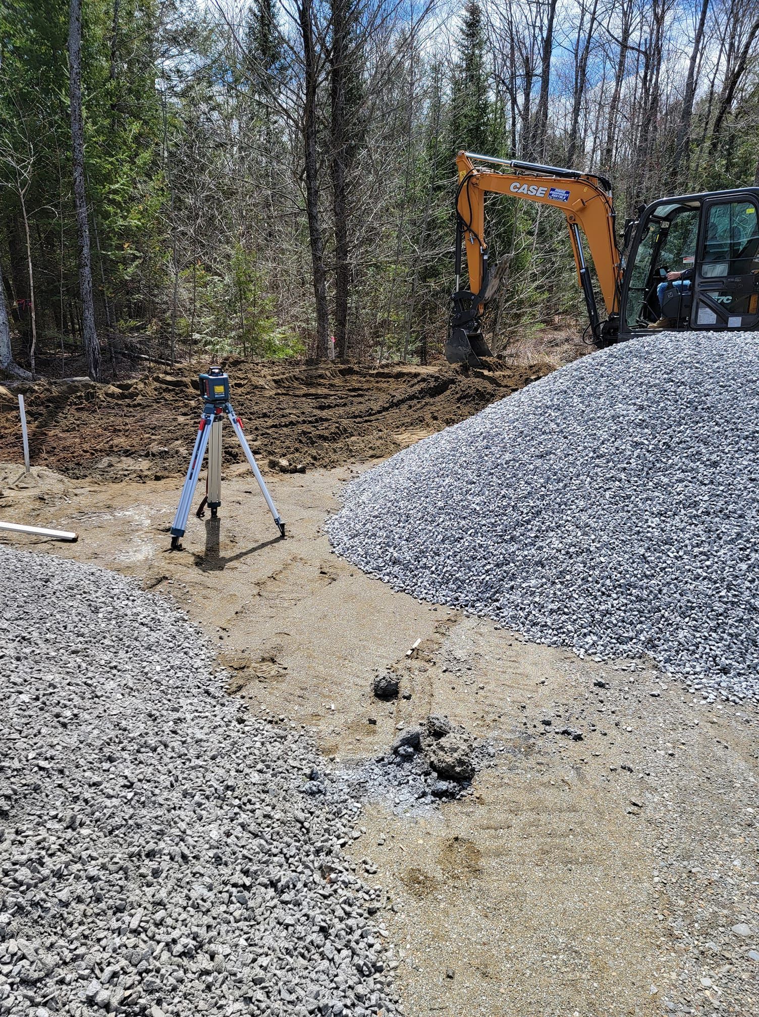 A surveyor’s tripod stands on a gravel surface next to piles of crushed stone with construction equipment in the background.
