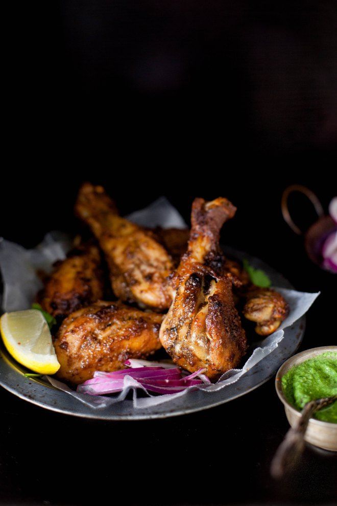 Grilled chicken drumsticks on a dark plate with lemon, red onion, and green chutney on a dark background