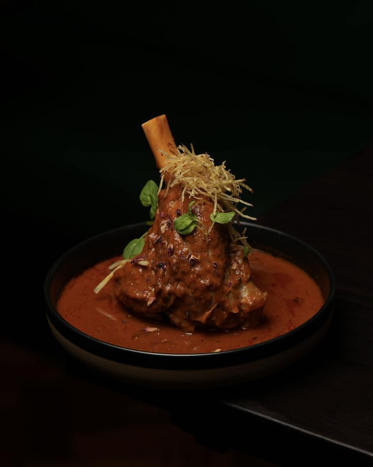 Braised lamb shank in red sauce, garnished with herbs, served on a dark plate.