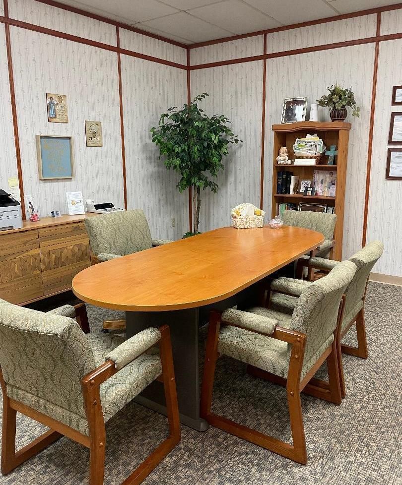 Conference room with oval wooden table, six chairs, cabinet, plant, and framed wall art.