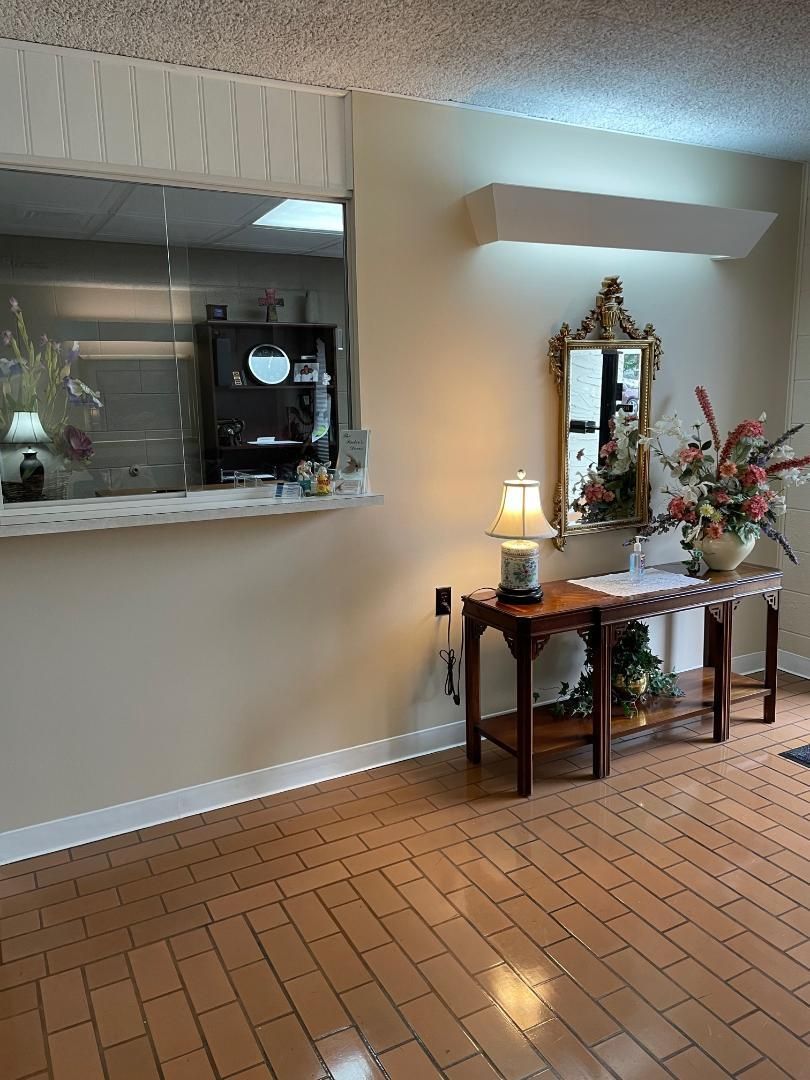 Lobby with a reception counter, wooden table, mirror, and floral arrangement on a tiled floor.