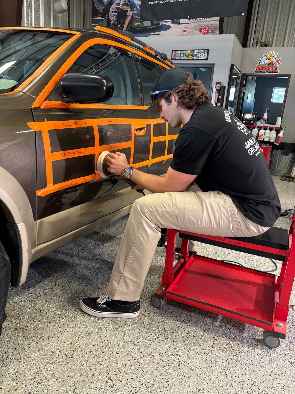 Man polishing a car door sectioned off with tape in a shop. He sits on a red stool, using a buffer.