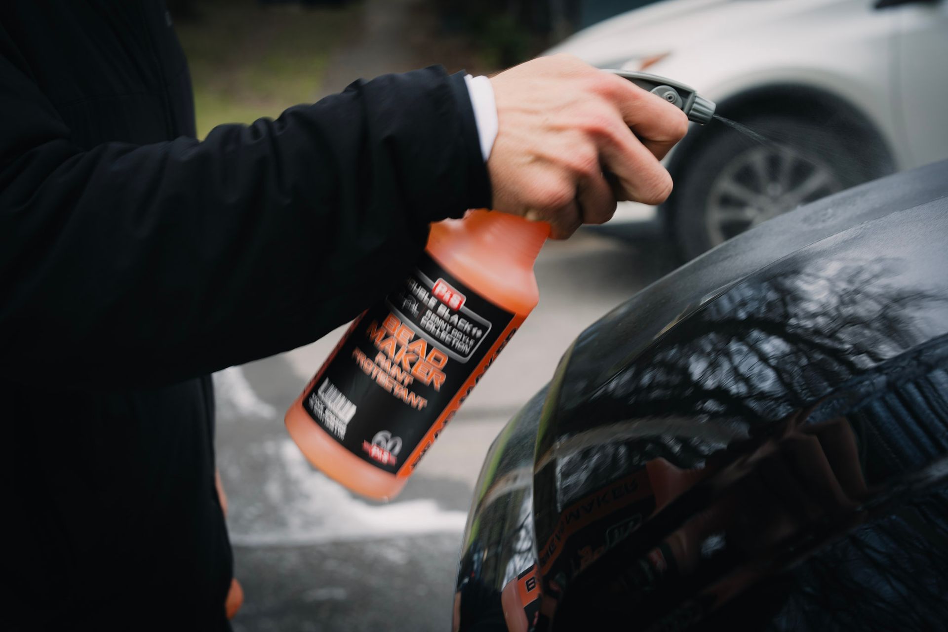 Person sprays car with orange bottle, presumably car wax or detailer; outdoor setting.