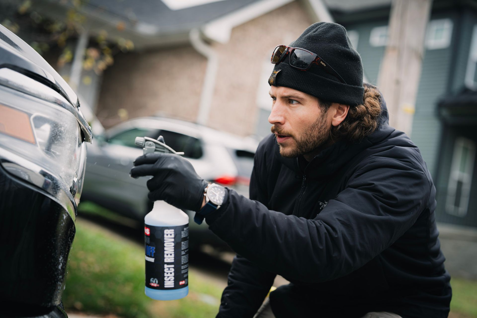 Man in black hat and gloves sprays car with bottle of product outdoors.