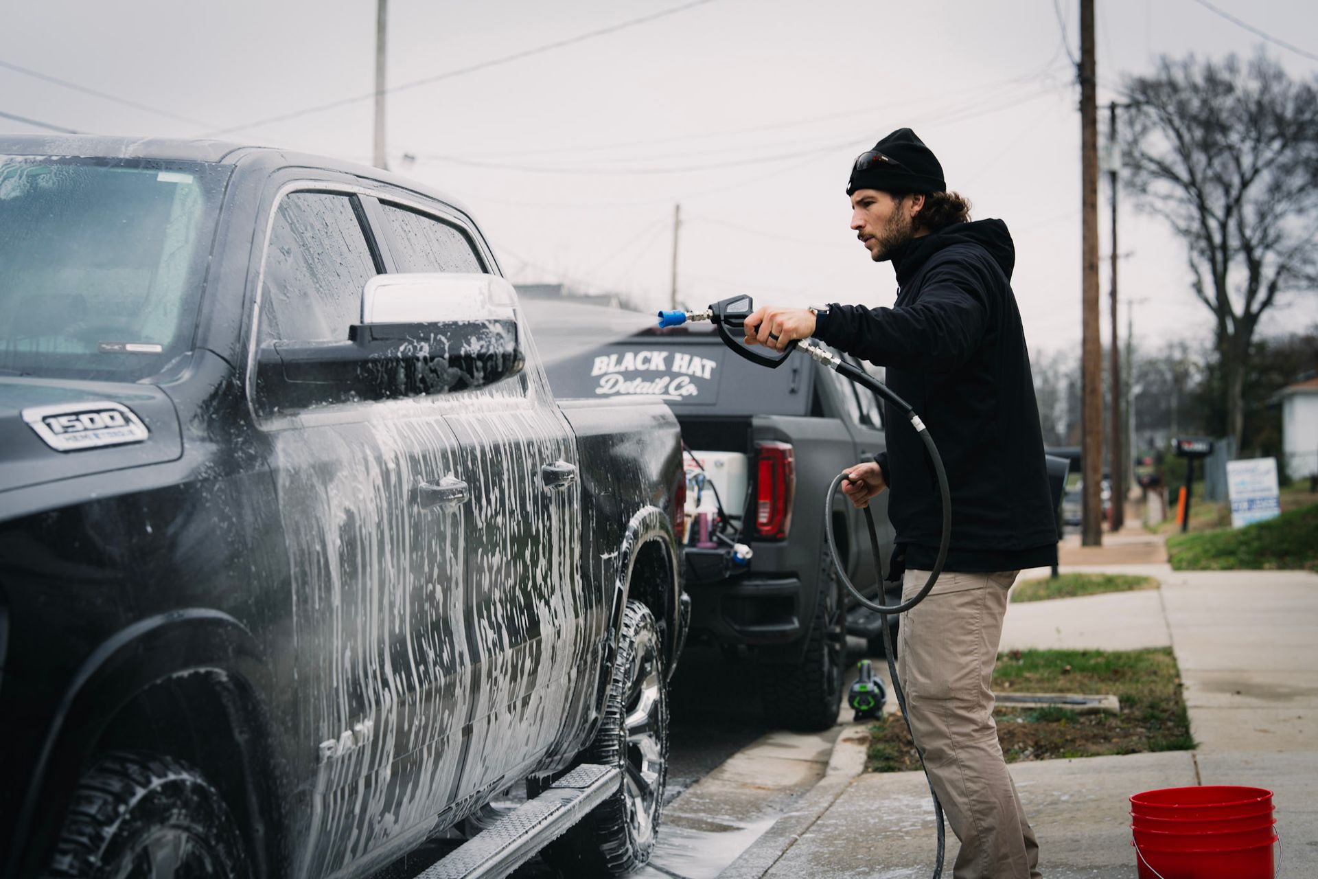 Man washes a black truck with a pressure washer outdoors; another truck nearby.