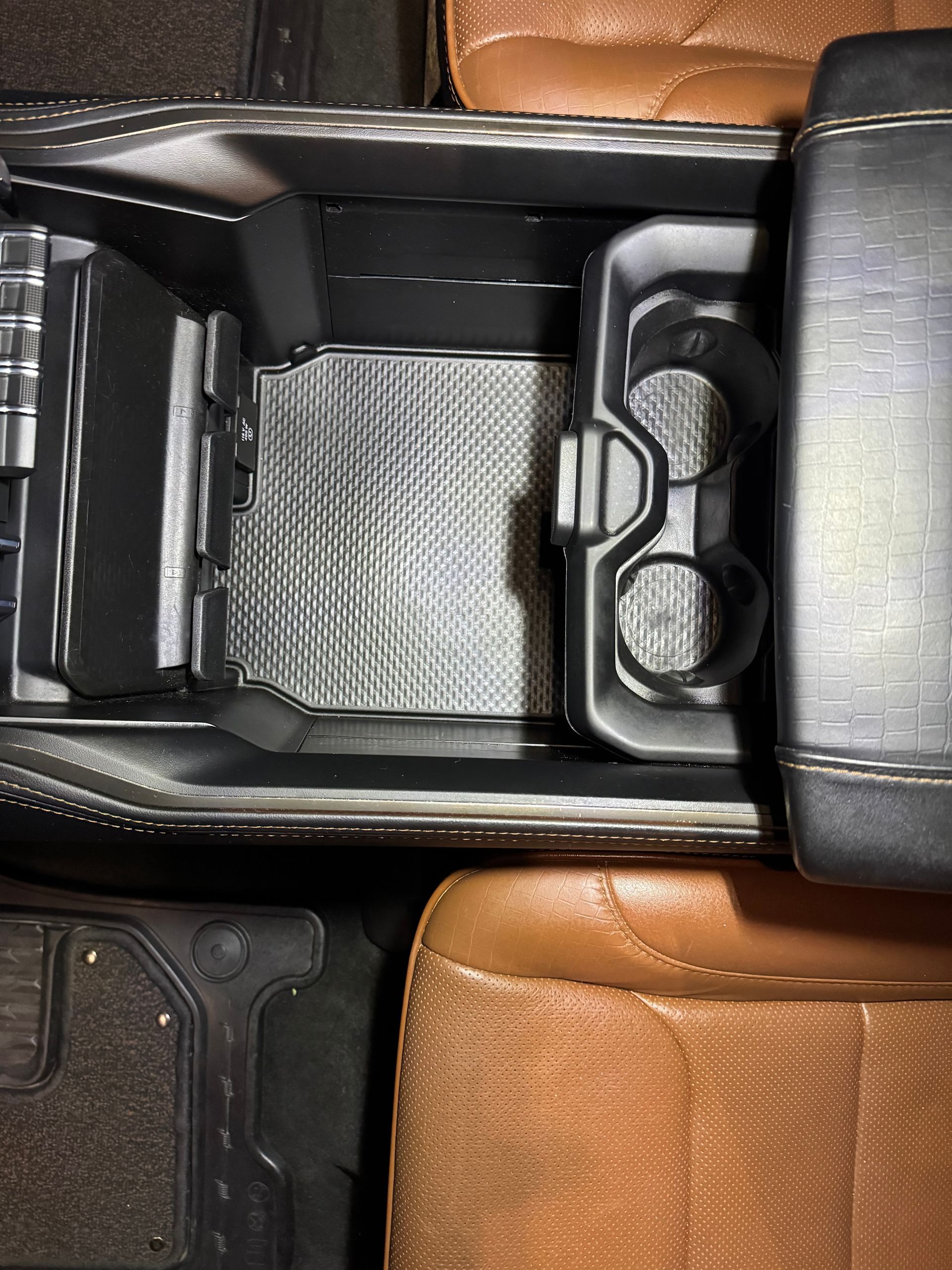 Open car console with cup holders, textured gray mat, and tan leather seats.