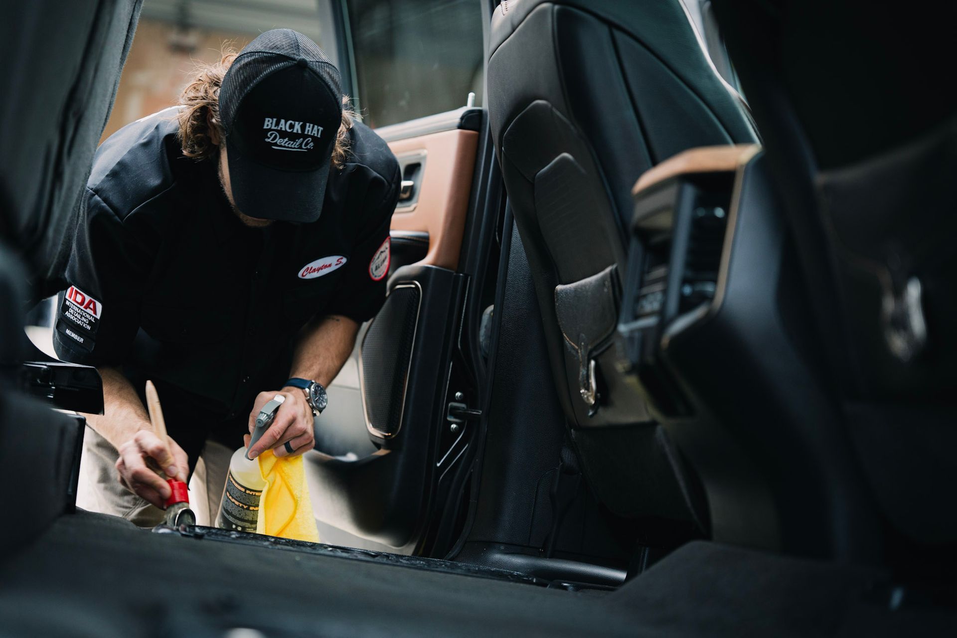 Person cleaning inside of a vehicle. Black clothing, cap, using spray bottle and tool. Gray and tan interior.