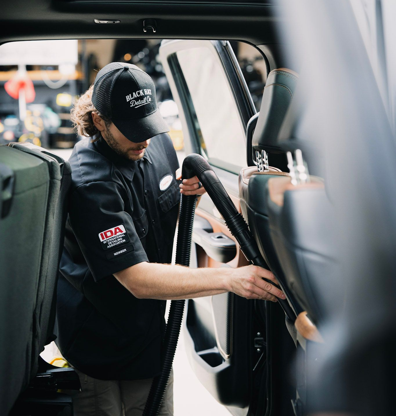 Person adjusts the seat inside a vehicle. Black shirt, cap, focused expression. Bright setting.