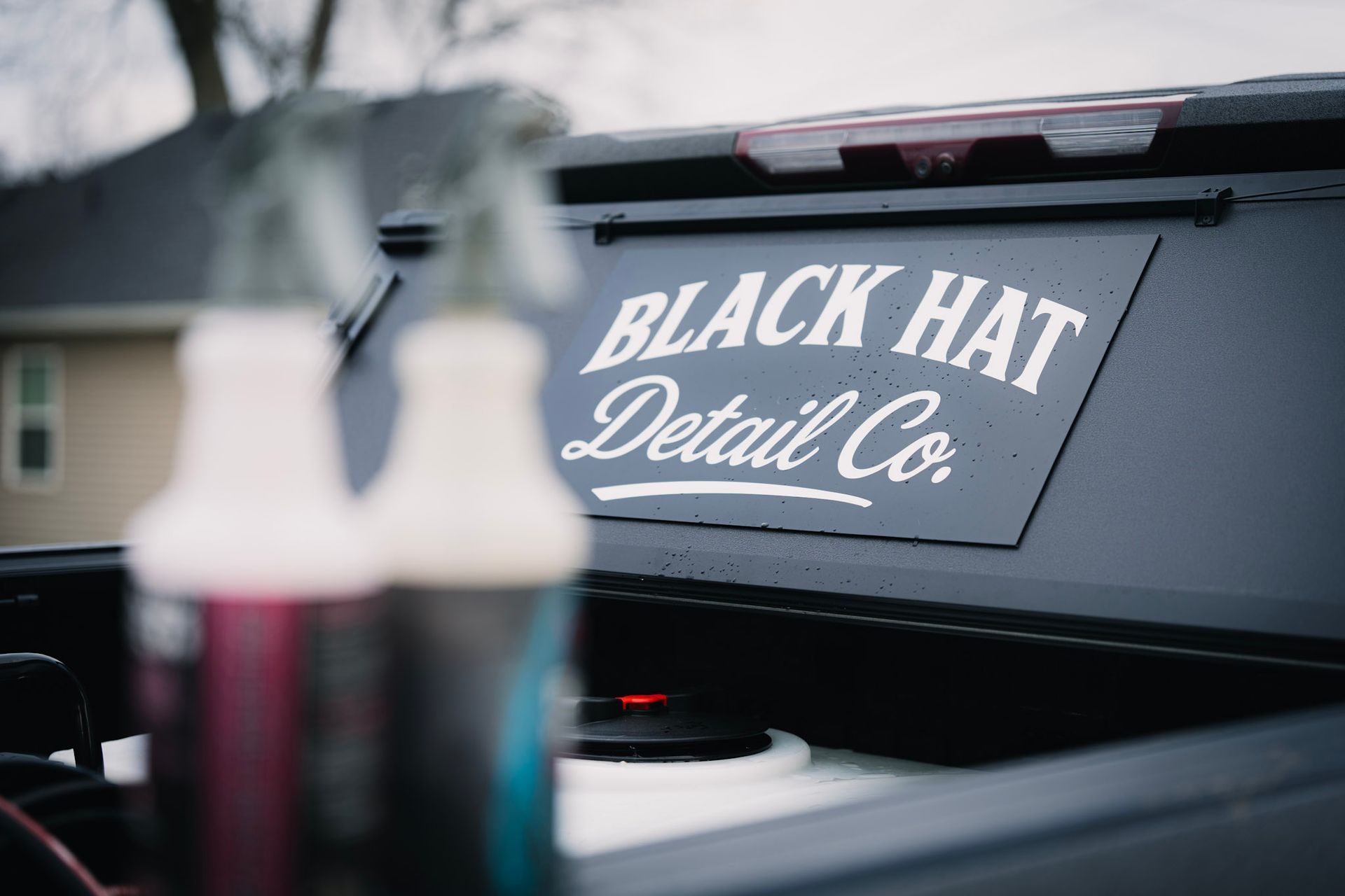 Black Hat Detail Co. logo on a black truck bed with blurred detail bottles in the foreground.