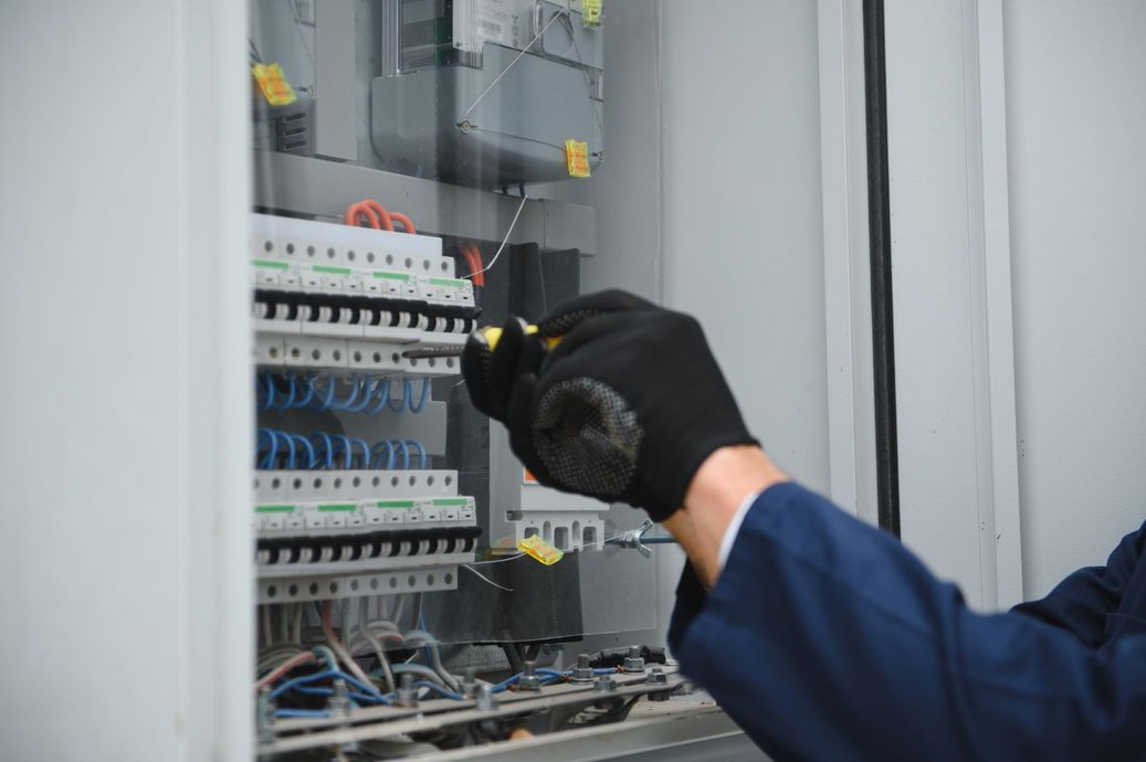 Electrician in a blue uniform, wearing black gloves, working on electrical panel wiring with a screwdriver.