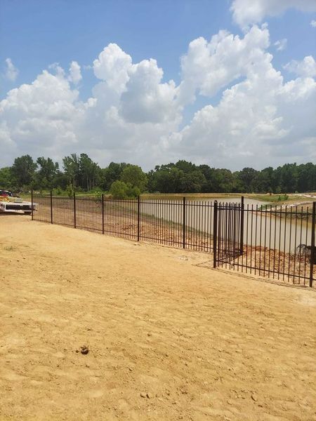 Dirt lot with a long metal fence under a cloudy blue sky, with trees in the background