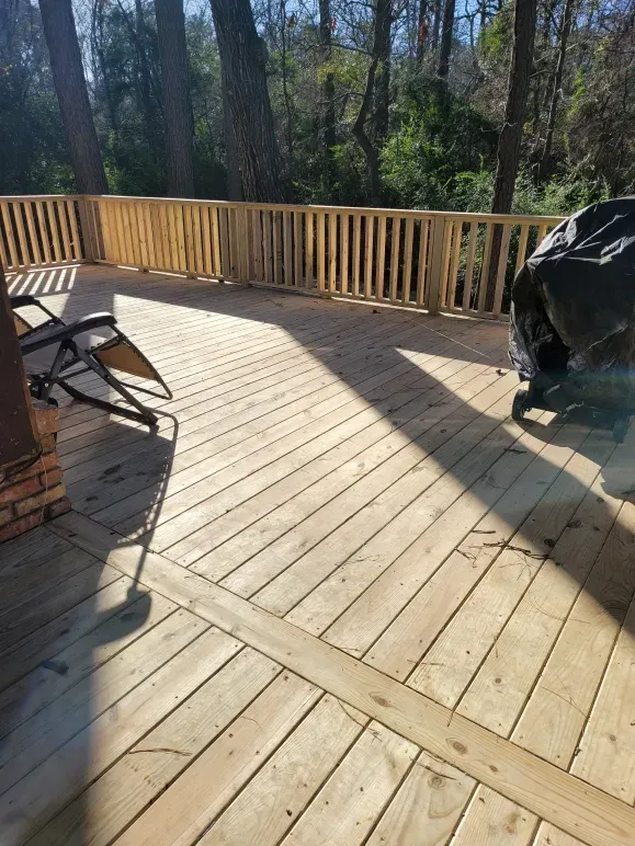 Sunlit wooden deck with railing, chair, and covered grill beside trees