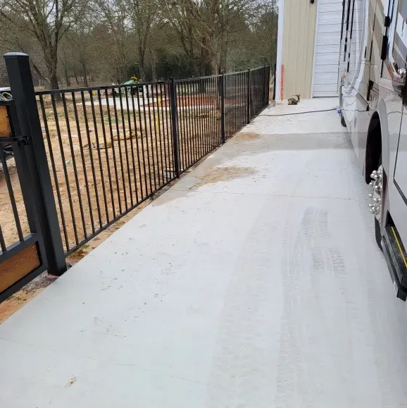 White RV parked beside a concrete walkway with black metal fencing along a wooded yard