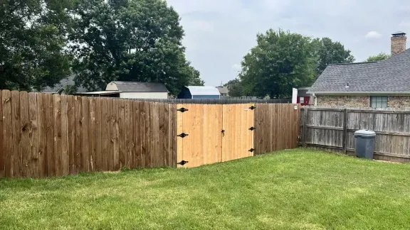 Backyard lawn with wooden fence and tan double gate, trees and a house visible behind.