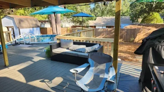 Backyard deck with patio furniture, blue umbrellas, and a covered grill in a sunny wooded setting