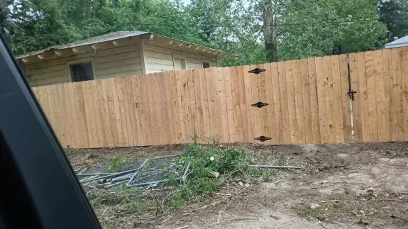 Wooden privacy fence beside a dirt yard, with a small shed and trees in the background.