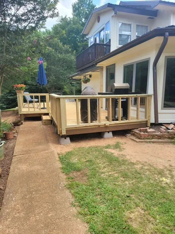 Backyard deck with light wood railing beside a house, bordering a concrete path and grassy yard