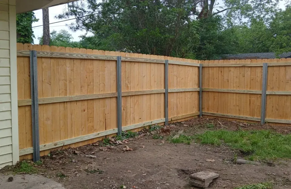 Wooden privacy fence around a bare backyard corner with a post and some trees beyond.