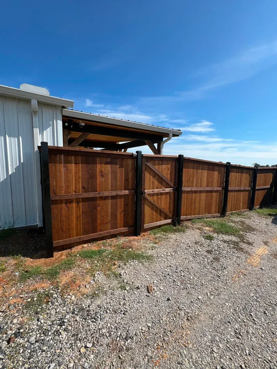 Wooden fence and gate beside a white metal building, with gravel ground and blue sky.