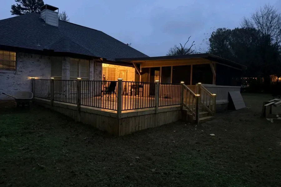 Twilight view of a lit wooden deck attached to a house, with railings and steps in a grassy yard