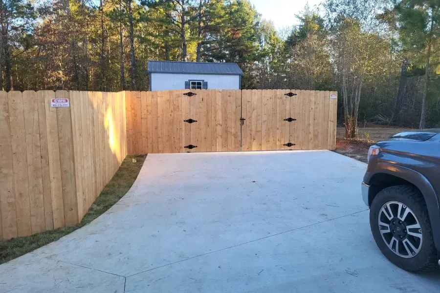 Wooden privacy fence around a concrete driveway, with a parked SUV at right and trees in the background.