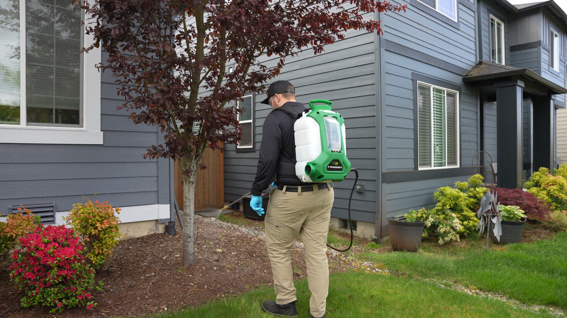 Person using a backpack sprayer treats plants beside a gray house and landscaped yard.